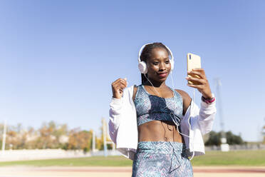 Sportlerin hört Musik, während sie ein Selfie mit ihrem Smartphone gegen den klaren Himmel macht - JCCMF00515