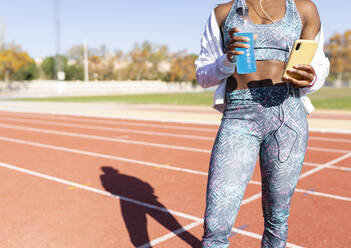Female sportsperson holding smart phone and energy drink while standing on running track - JCCMF00519