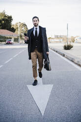 Businessman with briefcase and mobile phone walking on road - EBBF01990