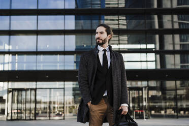 Entrepreneur with hands in pocket carrying briefcase while standing against modern building - EBBF01995