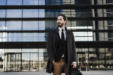 Unternehmer mit Händen in der Tasche, der eine Aktentasche trägt, während er vor einem modernen Gebäude steht - EBBF01995
