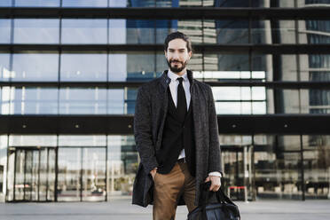 Männlicher Unternehmer mit Händen in der Tasche, der eine Aktentasche trägt, während er vor einem modernen Gebäude steht - EBBF01996