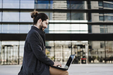 Young businessman working on laptop while sitting against building - EBBF01999