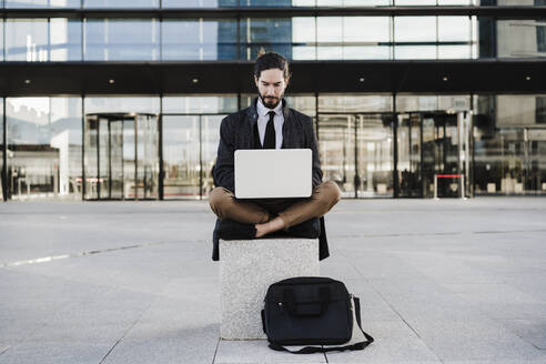 Unternehmer, der im Schneidersitz auf einer Bank sitzend am Laptop arbeitet - EBBF02001