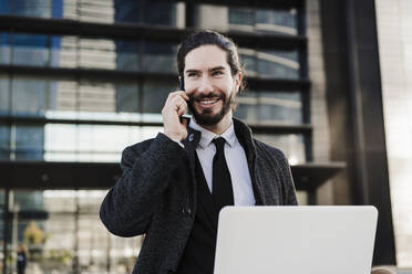 Smiling businessman with laptop talking on mobile phone while sitting outdoors - EBBF02002