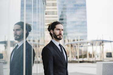 Young businessman wearing suit looking away while standing against glass wall - EBBF02011