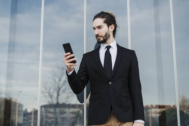 Geschäftsmann, der ein Smartphone benutzt, während er an einer Glaswand steht - EBBF02014