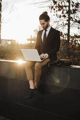 Geschäftsmann, der einen Laptop benutzt, während er bei Sonnenuntergang auf einer Stützmauer sitzt - EBBF02015