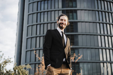 Junger Geschäftsmann mit Händen in den Taschen, der lächelnd vor einem Gebäude steht, lizenzfreies Stockfoto