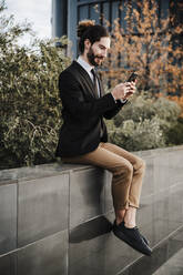 Geschäftsmann, der ein Mobiltelefon benutzt, während er auf einer Stützmauer sitzt - EBBF02021