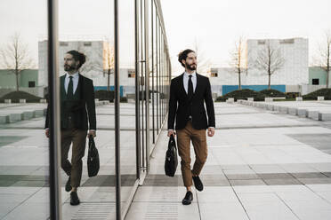 Young businessman with briefcase looking away while walking by glass wall - EBBF02029