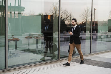 Young businessman using mobile phone while walking on footpath - EBBF02030