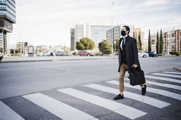 Geschäftsmann mit Jacke und Gesichtsmaske, der eine Aktentasche hält, während er auf einer Straße in der Stadt geht - EBBF02031