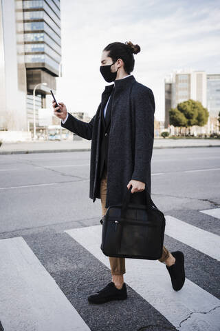 Young businessman wearing face mask using mobile phone while walking on road stock photo