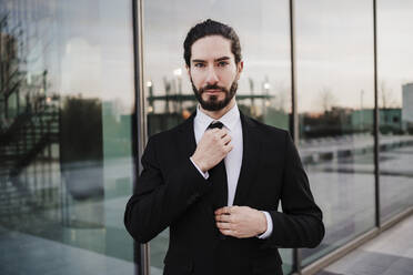 Businessman wearing suit adjusting tie while standing against glass wall - EBBF02041