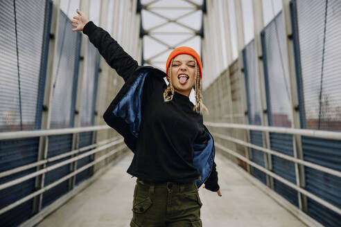 Happy young woman with eyes closed sticking out tongue while dancing on footbridge - ACPF00989