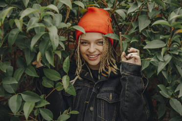 Lächelnde schöne Frau mit orangefarbener Strickmütze steht inmitten von Pflanzen im Park - ACPF01009
