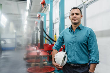 Smiling engineer with hands in pockets holding hardhat while standing in factory - DIGF14041