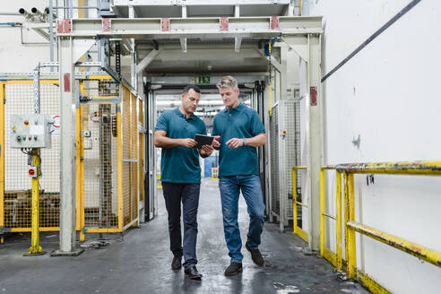 Mature male engineer colleagues walking while discussing over digital tablet in factory - DIGF14044
