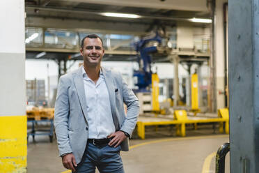 Smiling businessman with hand on hip standing in factory - DIGF14093