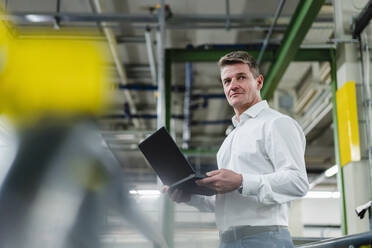 Lächelnder reifer Manager mit Laptop, der in einer Fabrik steht und vor sich hin träumt - DIGF14106