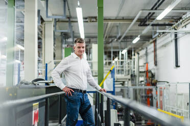 Lächelnder reifer Geschäftsmann mit Hand auf der Hüfte an einem Geländer in einer Fabrik stehend - DIGF14108