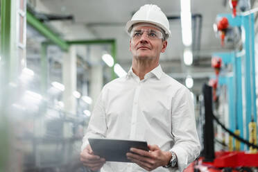 Älterer Geschäftsmann mit Schutzhelm, der ein digitales Tablet hält und in einer Fabrik wegschaut - DIGF14121