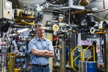 Male entrepreneur with arms crossed contemplating while standing against machinery in factory - DIGF14138