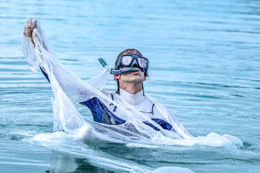 Mature man wearing scuba mask tearing plastic in sea - GGGF00736