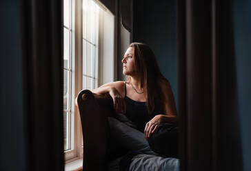 Spiegelreflexion einer hübschen Frau, die in einem dunklen Raum aus dem Fenster schaut. - CAVF91466