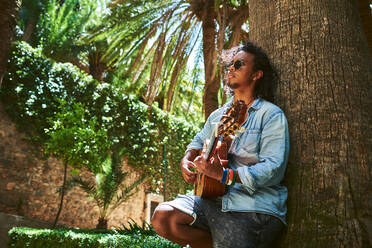 Junger Musiker mit klassischer Gitarre spielt im Schatten einiger Bäume in einem Park. - CAVF91469