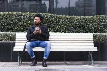 Mittlerer erwachsener Mann mit Afro-Haar, der ein Smartphone benutzt, während er auf einer Bank in der Stadt sitzt - PNAF00451