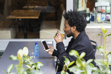 Mann mit Afro-Haar, der in einem Straßencafé ein Getränk zu sich nimmt und dabei ein Mobiltelefon benutzt - PNAF00456