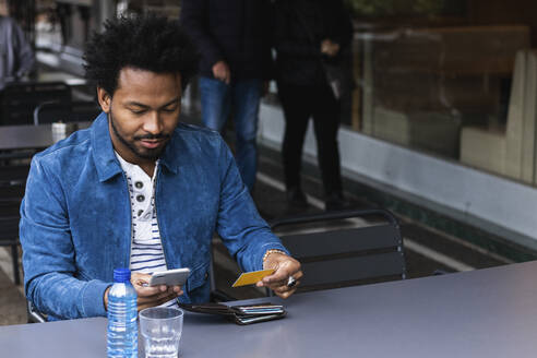 Mittlerer Erwachsener mit Kreditkarte und Smartphone zum Bezahlen in einem Straßencafé - PNAF00461