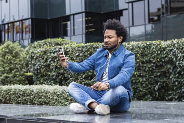 Mittlerer erwachsener Mann mit Afro-Haar, der ein Selfie über ein Smartphone macht, während er im Freien sitzt - PNAF00464