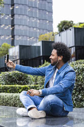 Lächelnder Mann mit Afro-Haar, der ein Selfie über ein Smartphone macht, während er an einem Gebäude sitzt - PNAF00465