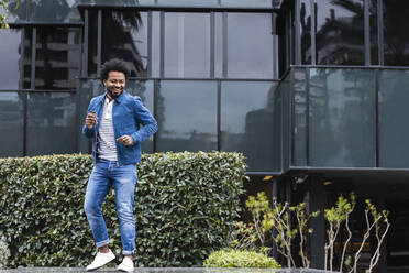 Cheerful man dancing while listening music against modern building in city - PNAF00466