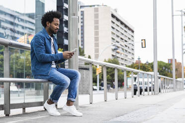 Stilvoller Mann mit Afro-Haar, der ein Mobiltelefon benutzt, während er auf einem Geländer in der Stadt sitzt - PNAF00468