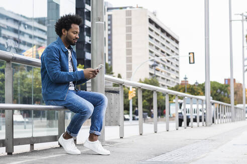 Stilvoller Mann mit Afro-Haar, der ein Mobiltelefon benutzt, während er auf einem Geländer in der Stadt sitzt - PNAF00468