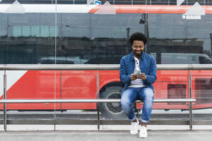 Lächelnder stilvoller Mann im mittleren Erwachsenenalter, der ein Mobiltelefon benutzt, während er an einer Bushaltestelle sitzt - PNAF00469