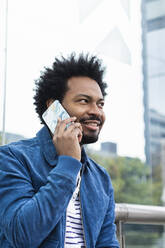 Nahaufnahme eines stilvollen Mannes mit Afro-Haar, der im Freien sitzend über ein Smartphone spricht - PNAF00470