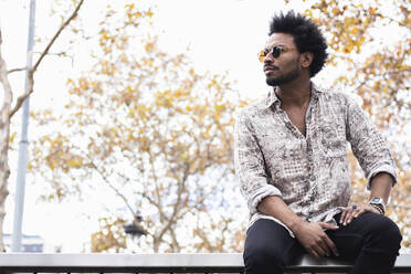 Stylish mid adult man with afro hair wearing sunglasses sitting on railing - PNAF00486