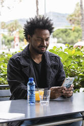 Gut aussehender Mann mit Afro-Haar, der ein Mobiltelefon benutzt, während er in einem Straßencafé sitzt - PNAF00490