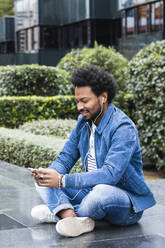 Stylish man listening music and using smart phone while sitting on retaining wall - PNAF00494