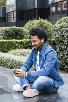 Stilvoller Mann, der Musik hört und sein Smartphone benutzt, während er auf einer Stützmauer sitzt - PNAF00494