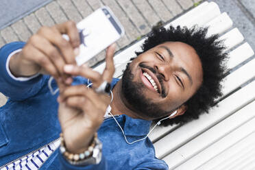 Nahaufnahme eines fröhlichen Mannes mit Afro-Haar, der auf einer Bank liegend Musik hört und ein Mobiltelefon benutzt - PNAF00495