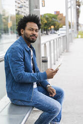 Selbstbewusster Mann mit Afro-Haar, der ein Mobiltelefon benutzt, während er auf einem Sitz an einer Bushaltestelle sitzt - PNAF00497