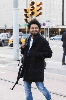 Lächelnder Mann mit Afro-Haar, der beim Gehen auf der Straße in der Stadt wegschaut - PNAF00499