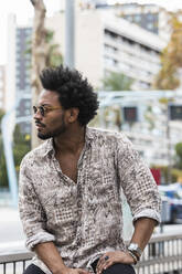 Stilvoller erwachsener Mann mit Afro-Haar, der eine Sonnenbrille trägt, während er in der Stadt sitzt - PNAF00507