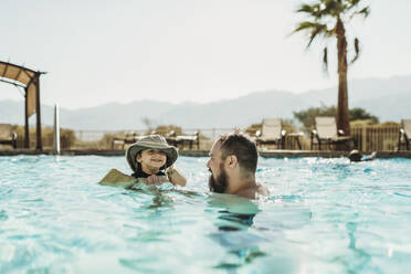 Father and toddler son swimming in pool with mountains and palm trees - CAVF91501